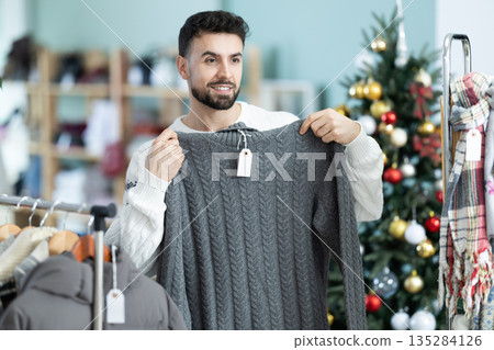 man chooses a sweater against the background of a Christmas tree in a store man chooses a sweater against the background of a Christmas tree in a store 135284126