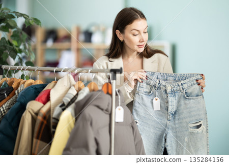 Happy young female client carefully choosing stylish jeans trousers in clothing store. Shopping and seasonal sales concept Happy young female client carefully choosing stylish jeans trousers in clothing store. Shopping and seasonal sales concept 135284156