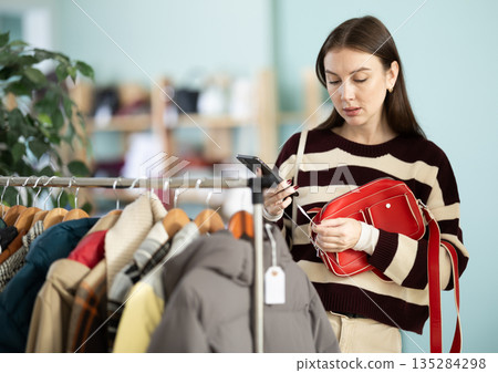 Positive young woman customer standing in interior of clothing boutique and choosing fashionable handbag, scanning barcode with smartphone to get information Positive young woman customer standing in interior of clothing boutique and choosing fashionable handbag, scanning barcode with smartphone to get information 135284298
