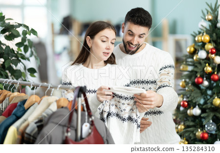 Married couple choose sweaters against background of Christmas tree in clothing store Married couple choose sweaters against background of Christmas tree in clothing store 135284310
