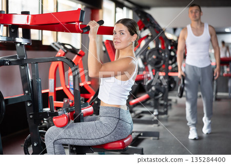 Girl doing strength training in gym, exercising upper body muscles on shoulder press machine 135284400