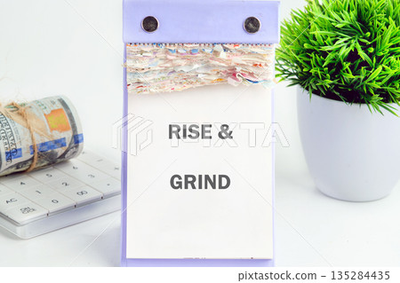 The phrase RISE and GRIND is seen on a tear-off calendar sitting on a white desk. Next to the calendar is a roll of Dollars, keyboard, and green plant 135284435