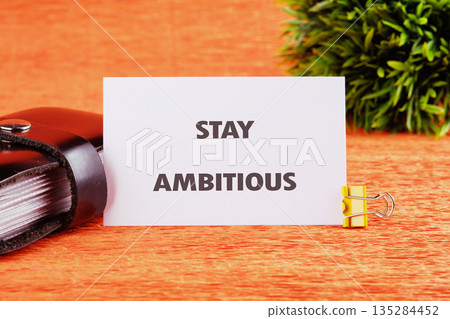 A card displays the phrase STAY AMBITIOUS, clipped with a golden binder clip. It sits near a dark book and a green plant, all on a bright orange surface, evoking motivation A card displays the phrase STAY AMBITIOUS, clipped with a golden binder clip. It sits near a dark book and a green plant, all on a bright orange surface, evoking motivation 135284452