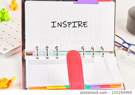 An open planner reads the word inspire, sitting amid crumpled paper, a calculator, glasses, and other desk items on a white surface, creating an atmosphere of inspiration 135284469