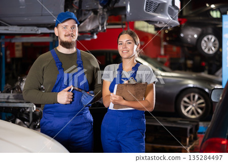 Portrait of two auto mechanics - man and girl against the background of cars in car service center Portrait of two auto mechanics - man and girl against the background of cars in car service center 135284497