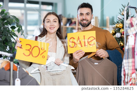 Joyful couple holding sales poster in clothing store Joyful couple holding sales poster in clothing store 135284625