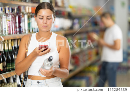 Young female buyer scan QR code on bottle of vodka 135284889