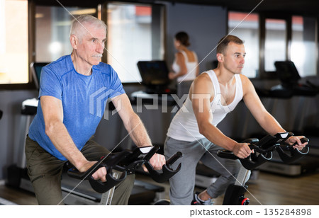 Two men exercising on stationary exercise bikes in gym 135284898