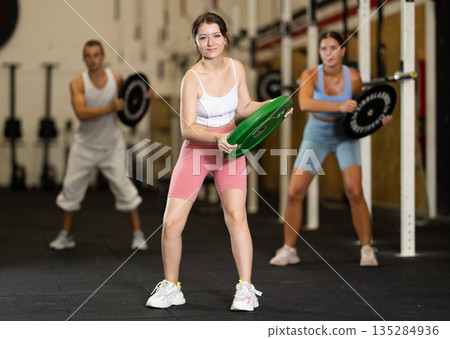 Closeup of athletic sports girl standing with plate of barbell in hands in fitness studio 135284936