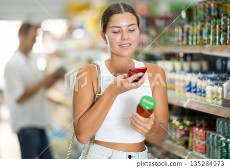Girl scanning barcode or qr code with smartphone on sauce label in supermarket 135284973