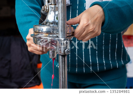 A woman extracts pomegranate juice using a manual juicer. She has dark hair and wears a blue outfit. The scene is set in a market environment. 135284981