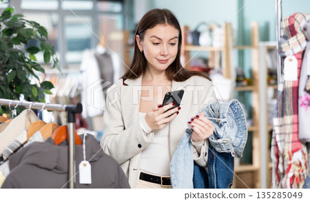 Happy female client carefully choosing stylish jeans trousers in clothing store and scanning barcode with smartphone 135285049