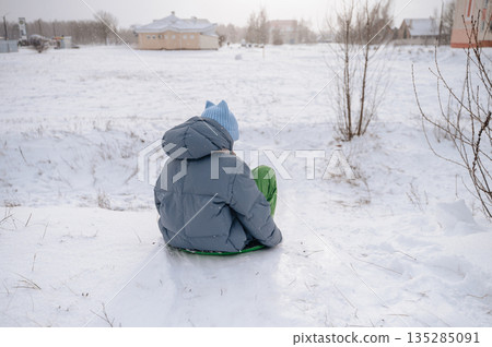 Girl in winter jacket sliding down snowy hill, playful childhood fun outdoors 135285091