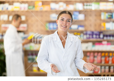 At pharmacy, girl apothecary waiting for customers, invite with gesture go to department of cosmetic At pharmacy, girl apothecary waiting for customers, invite with gesture go to department of cosmetic 135285173