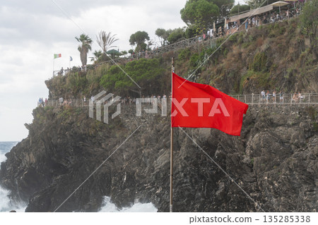 Red warning flag on a rocky seaside cliff above the sea. Concept of danger, storm, safety warning, strong waves and coastal conditions Red warning flag on a rocky seaside cliff above the sea. Concept of danger, storm, safety warning, strong waves and coastal conditions 135285338
