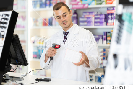Man pharmacist in cash area works with computer and goods, scans drugs, checks availability Man pharmacist in cash area works with computer and goods, scans drugs, checks availability 135285361