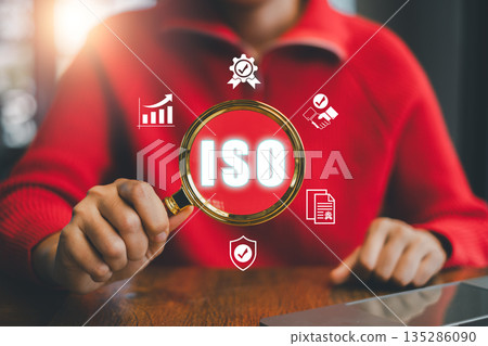 Business professional examines ISO certification with magnifying glass, surrounded by digital icons representing quality and standards. 135286090