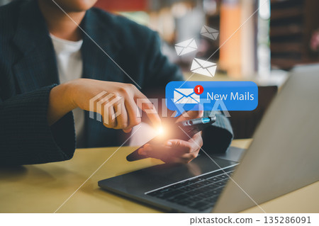A person using a smartphone to check new emails, with a laptop on the desk in a modern office environment. A person using a smartphone to check new emails, with a laptop on the desk in a modern office environment. 135286091