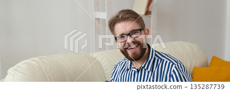 Portrait smiling man sitting on sofa in living room interior. Banner with copy space. Friendly mood, personal presence, and relaxed lifestyle portrait inside modern home. Portrait smiling man sitting on sofa in living room interior. Banner with copy space. Friendly mood, personal presence, and relaxed lifestyle portrait inside modern home. 135287739