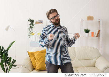 Man dancing while listening to music with headphones in living room. Personal entertainment, audio enjoyment, and expressive movement inside home interior. Man dancing while listening to music with headphones in living room. Personal entertainment, audio enjoyment, and expressive movement inside home interior. 135287776