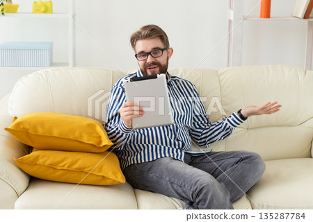 Man talking on video call using tablet while sitting on sofa. Remote communication, online meeting, and expressive conversation at home. Man talking on video call using tablet while sitting on sofa. Remote communication, online meeting, and expressive conversation at home. 135287784