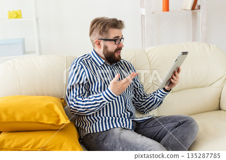 Man talking on video call using tablet while sitting on sofa. Remote communication, online meeting, and expressive conversation at home. Man talking on video call using tablet while sitting on sofa. Remote communication, online meeting, and expressive conversation at home. 135287785