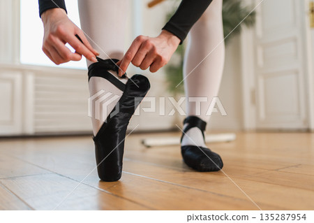 Ballerina puts on pointe shoes on leg in dance class. Classical ballet dancer woman in dance studio. Ballerina putting on tying classical pointe shoes for dance training. Ballet school concept Ballerina puts on pointe shoes on leg in dance class. Classical ballet dancer woman in dance studio. Ballerina putting on tying classical pointe shoes for dance training. Ballet school concept 135287954