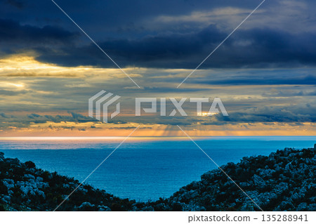 Serene Sunset Over Calm Sea With Rocky Hills Framing The Distant Golden Horizon 135288941