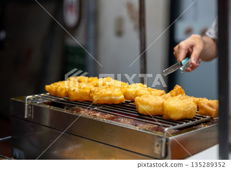 Close-up view of a street food cook turning freshly fried patongo dough with metal tongs, capturing the hot oil, golden texture, and authentic Thai street cuisine atmosphere. Close-up view of a street food cook turning freshly fried patongo dough with metal tongs, capturing the hot oil, golden texture, and authentic Thai street cuisine atmosphere. 135289352