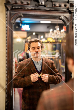 Chic man buyer fitting on classy wool blazer with checkered pattern in front of mirror in store. Chic man buyer fitting on classy wool blazer with checkered pattern in front of mirror in store. 135290258