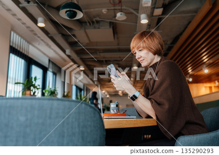 Mature female sitting at communal table and checking messages on smartphone. Modern workspace, adult independence, realistic scene of everyday digital business communication. 135290293