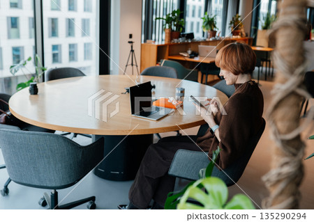 Middle age female with short red hair sitting at large round table and using smartphone next to open laptop. Everyday work, modern interior with plants, realistic adult lifestyle without urgency. 135290294