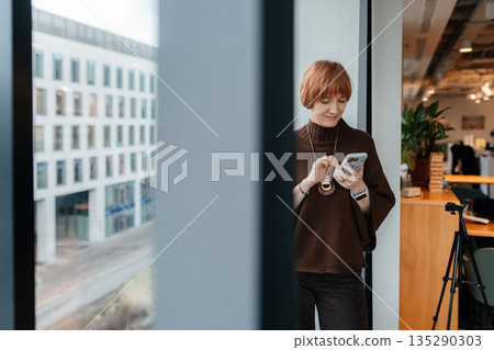 Focused female entrepreneur standing near large window in modern office, holding smartphone and reading information. Calm expression, business work environment, architectural background. 135290303