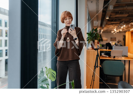 Middle age red haired woman wearing brown sweater, near open office and sending voice message on smartphone. Manager, consultant or content creator using audio communication, flexible digital work. Middle age red haired woman wearing brown sweater, near open office and sending voice message on smartphone. Manager, consultant or content creator using audio communication, flexible digital work. 135290307