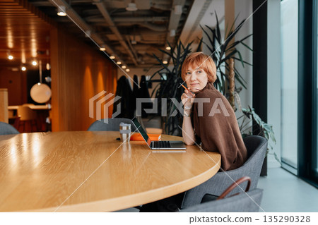 Cheerful professional woman freelancer holds glasses, wear brown sweater sitting at around desk with laptop, notepad and coffee cup. Cheerful professional woman freelancer holds glasses, wear brown sweater sitting at around desk with laptop, notepad and coffee cup. 135290328