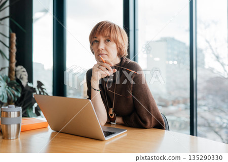 Stylish business woman sitting at wooden conference table, working on laptop and thinking, looking at camera in modern office interior with large window on background Stylish business woman sitting at wooden conference table, working on laptop and thinking, looking at camera in modern office interior with large window on background 135290330