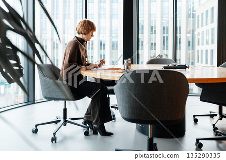 White woman aged 49 sitting alone at large conference table in modern office, working on laptop with notes and drink on table. Business professional planning meeting with large window. 135290335