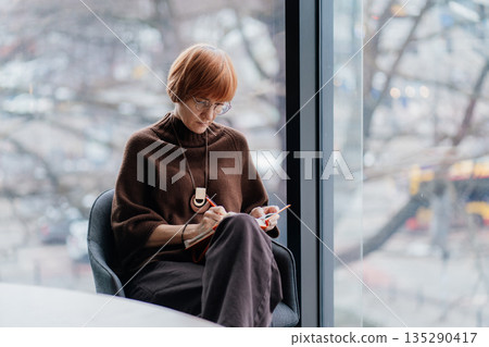 Experienced consultant or businesswoman sitting near large window and writing notes in planner. Concept of work, financial strategy, professional life, productivity, economy, modern workspace 135290417
