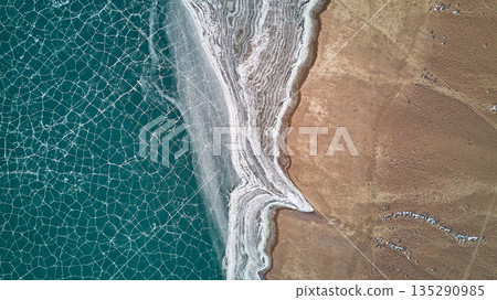 Aerial view of cracked ice on lake next to frozen shore and brown land Aerial view of cracked ice on lake next to frozen shore and brown land 135290985