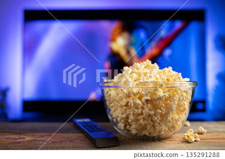 A glass bowl of popcorn and remote control in the background the TV works. Evening cozy watching a movie or TV series at home A glass bowl of popcorn and remote control in the background the TV works. Evening cozy watching a movie or TV series at home 135291288