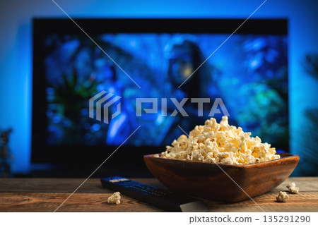 A wooden bowl of popcorn and remote control in the background the TV works. Evening cozy watching a movie or TV series at home A wooden bowl of popcorn and remote control in the background the TV works. Evening cozy watching a movie or TV series at home 135291290