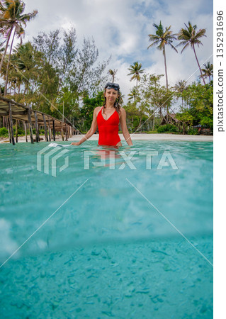 Woman in red swimsuit standing in clear tropical water with palm trees and beach resort behind Woman in red swimsuit standing in clear tropical water with palm trees and beach resort behind 135291696