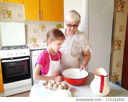 A grandmother and granddaughter stand at a table with a bowl, eggs, and a pitcher. A grandmother and granddaughter stand at a table with a bowl, eggs, and a pitcher. 135292691