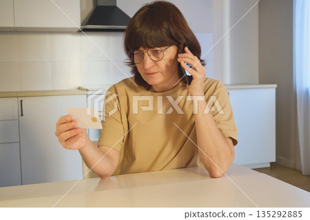 Woman holding a credit card and talking on a mobile phone in a kitchen at home Woman holding a credit card and talking on a mobile phone in a kitchen at home 135292885