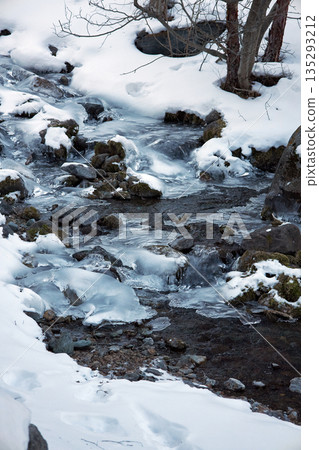 Frozen mountain stream covered in ice flowing through snowy landscape 135293212