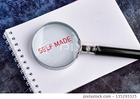 A magnifying glass is placed on a white, spiral notebook, showing the words Self Made in red. The background is dark and textured, creating a contrast A magnifying glass is placed on a white, spiral notebook, showing the words Self Made in red. The background is dark and textured, creating a contrast 135293325