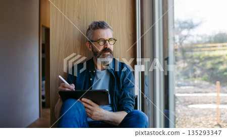 Man standing by the window with tablet and writing notes. Man standing by the window with tablet and writing notes. 135293347