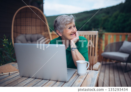 Mature woman sitting on balcony with laptop, working remotely. 135293434