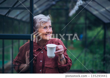 Senior woman standing in greenhouse and drinking coffee. 135293441