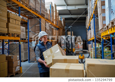 Older male warehouse worker pulling a pallet truck. Older male warehouse worker pulling a pallet truck. 135294799